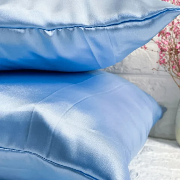 Fronha de cetim azul clara em almofadas empilhadas com fundo branco e flores rosa.